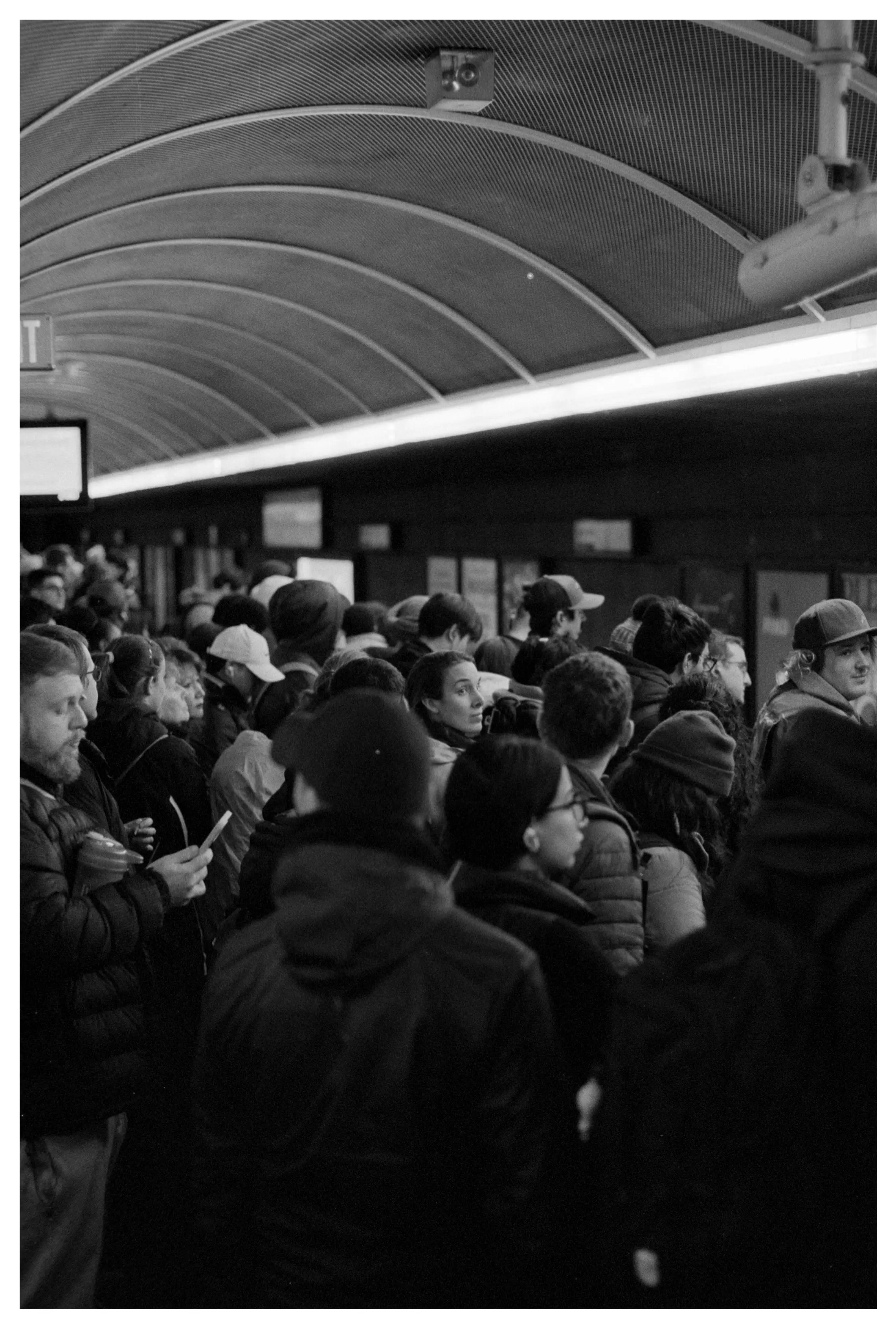 Crowded subway platform with people waiting under a curved ceiling with overhead lights.