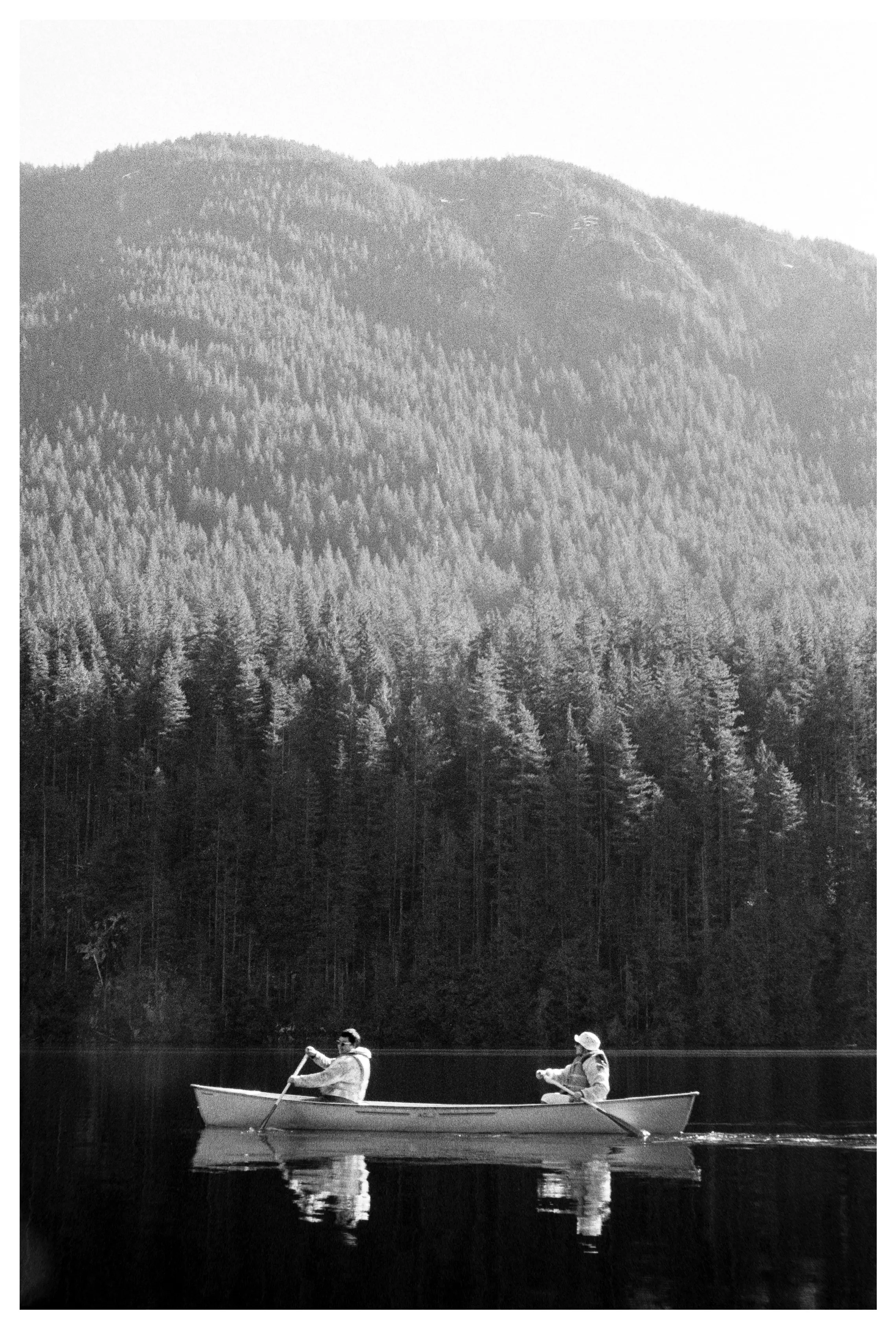 Two people rowing a canoe on a calm lake with a dense pine forest and mountain in the background.