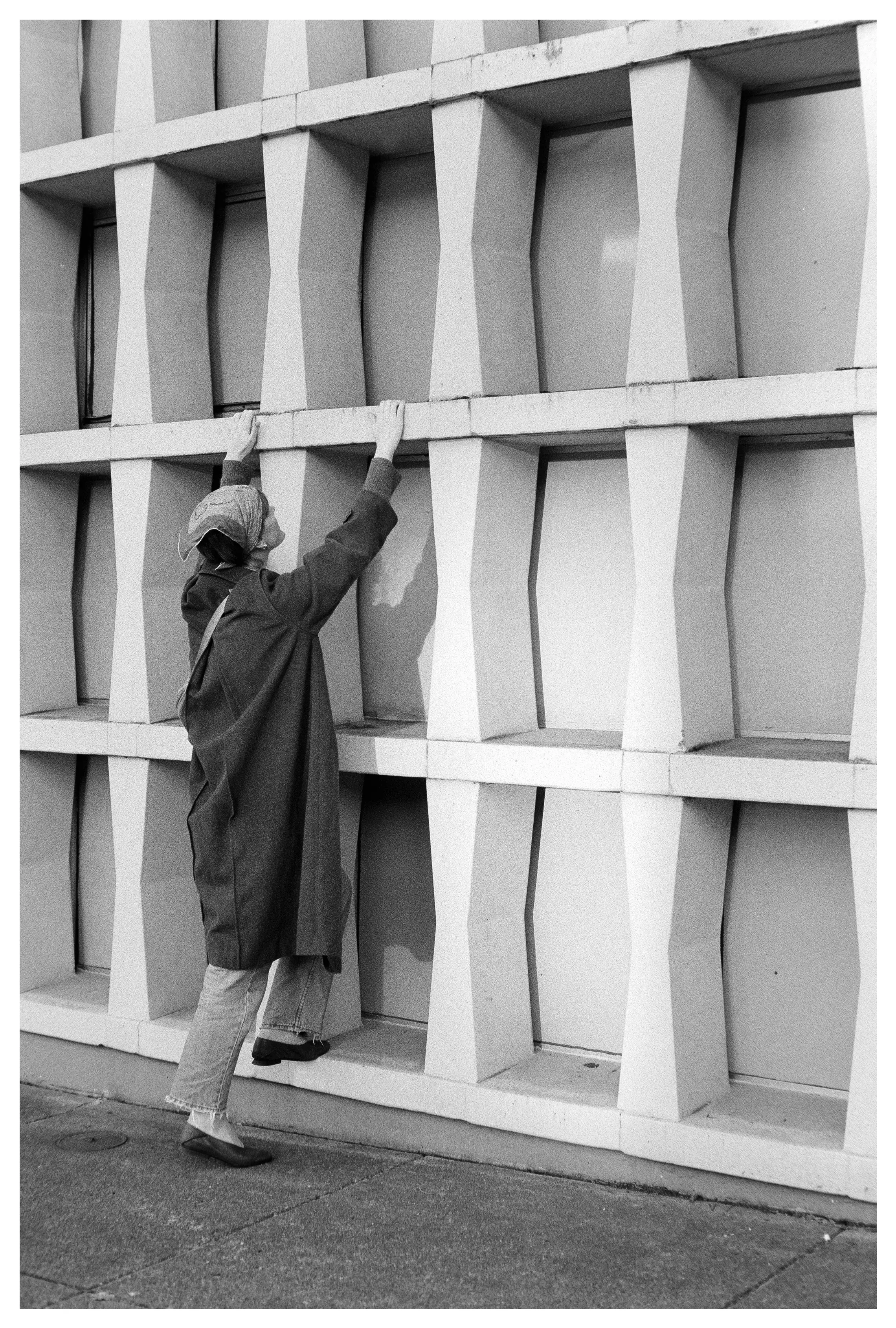 Person wearing a headscarf and long coat climbing a geometric concrete wall.