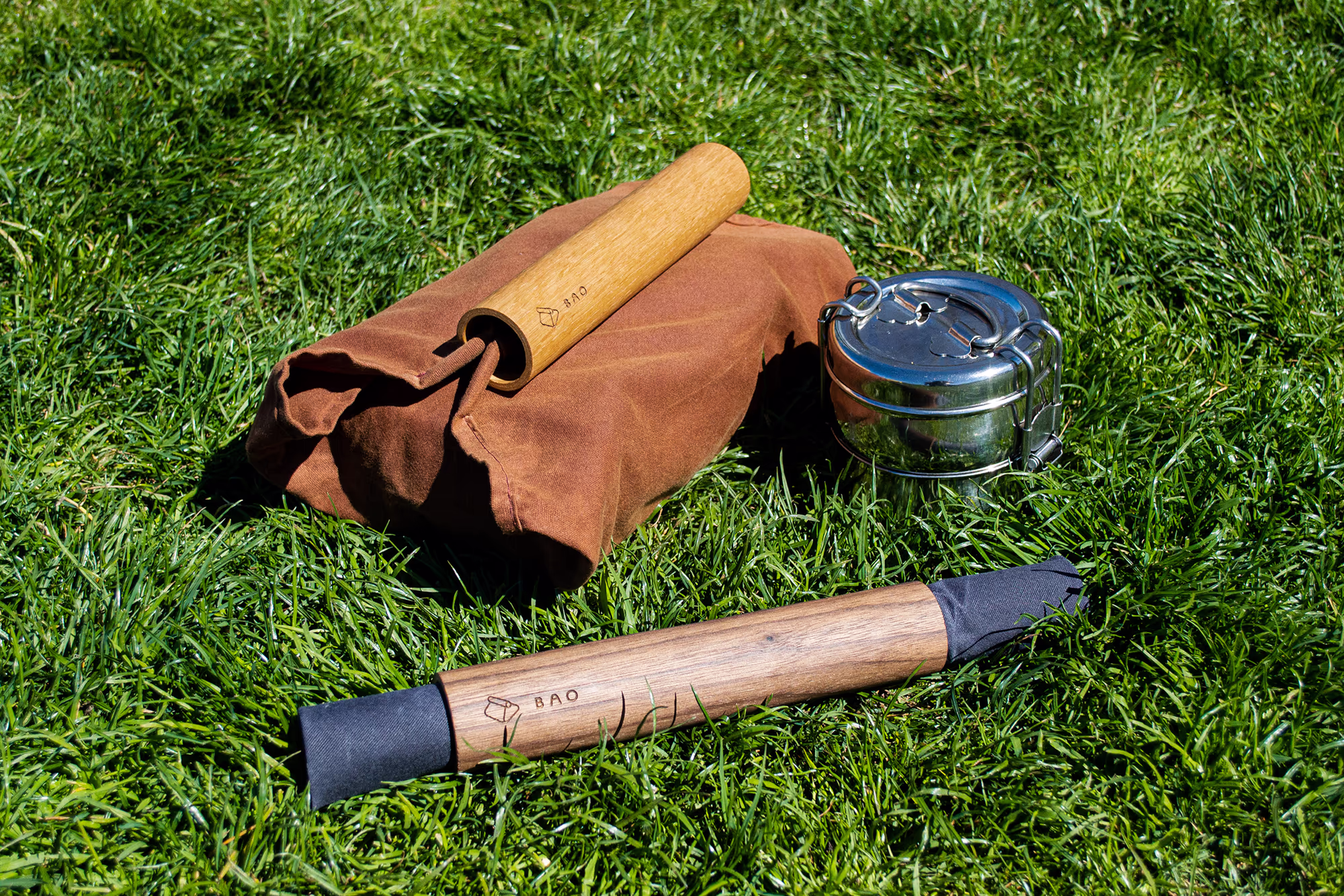 Brown fabric bag with a wooden handle, a stainless steel tiffin container, and a wooden cylindrical item with black fabric ends lying on green grass.