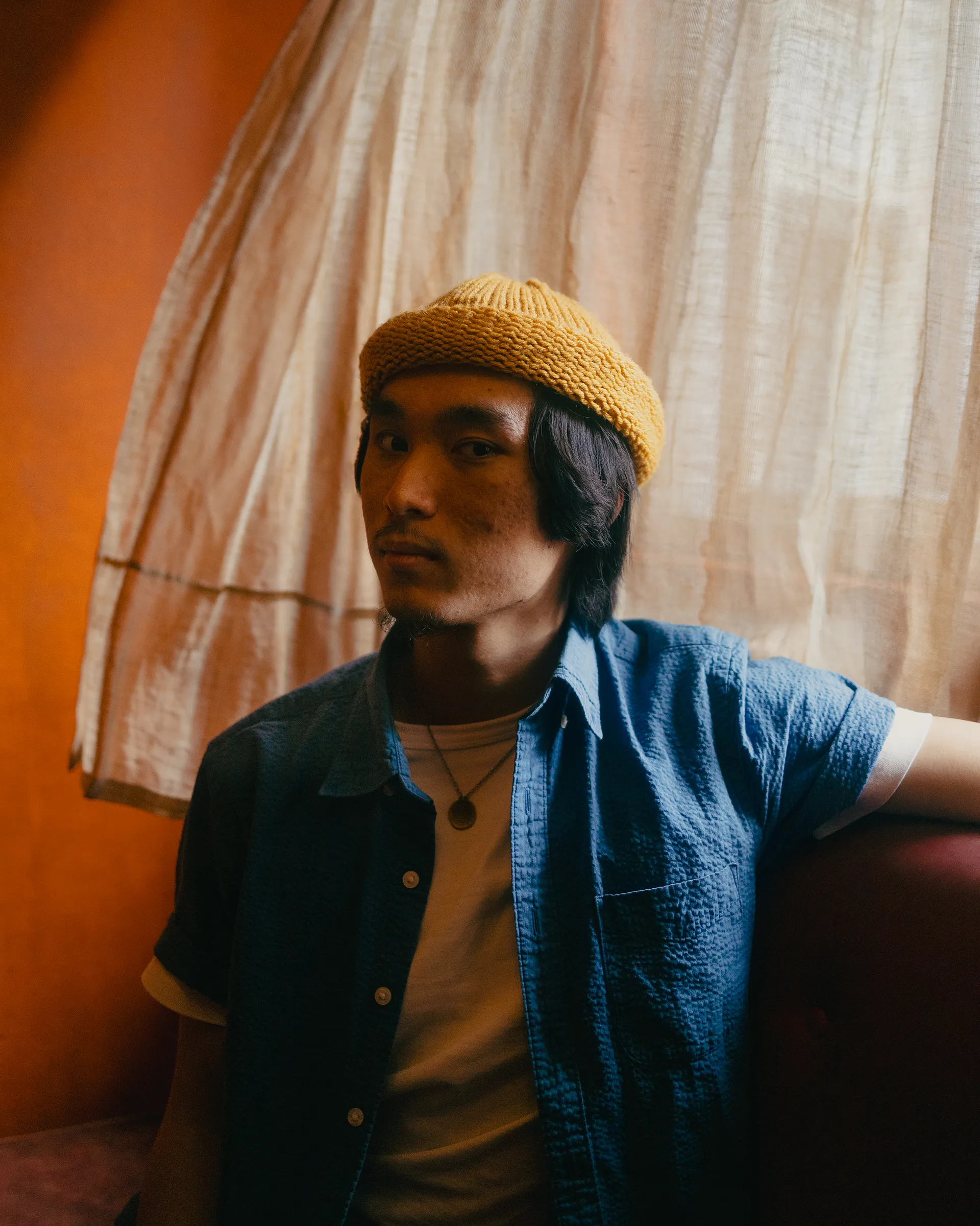 Young man wearing a yellow knit hat and blue shirt sitting indoors against a sheer curtain and orange wall.