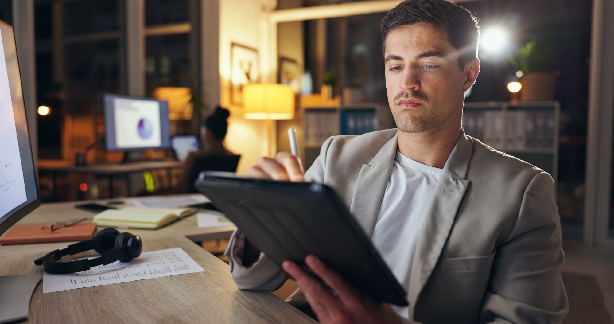 Man working late in an office, using a digital tablet at his desk with documents, headphones, and a computer monitor nearby. He is signing a contract with signeer by mesoneer.