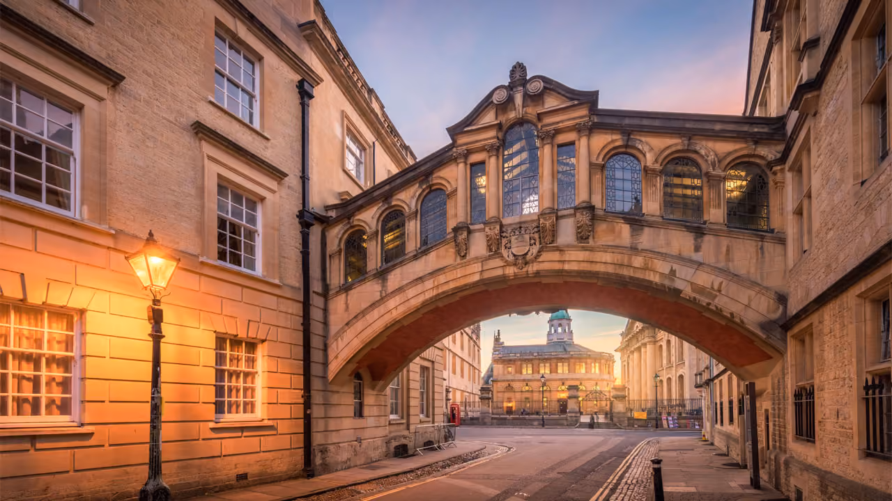 Historic arched stone bridge connecting buildings over a street lit by a lamp at sunset.