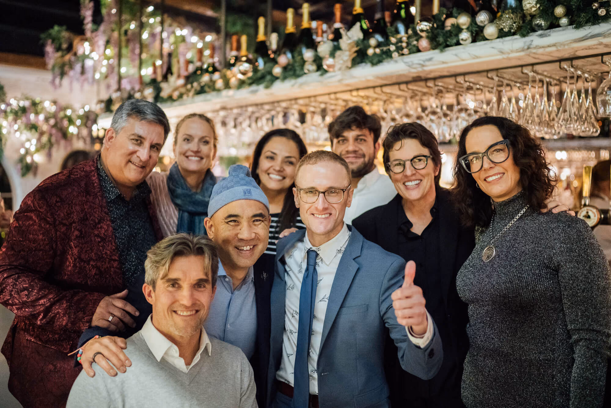 Group of smiling adults posing together in a warmly lit bar with festive decorations and hanging glasses.