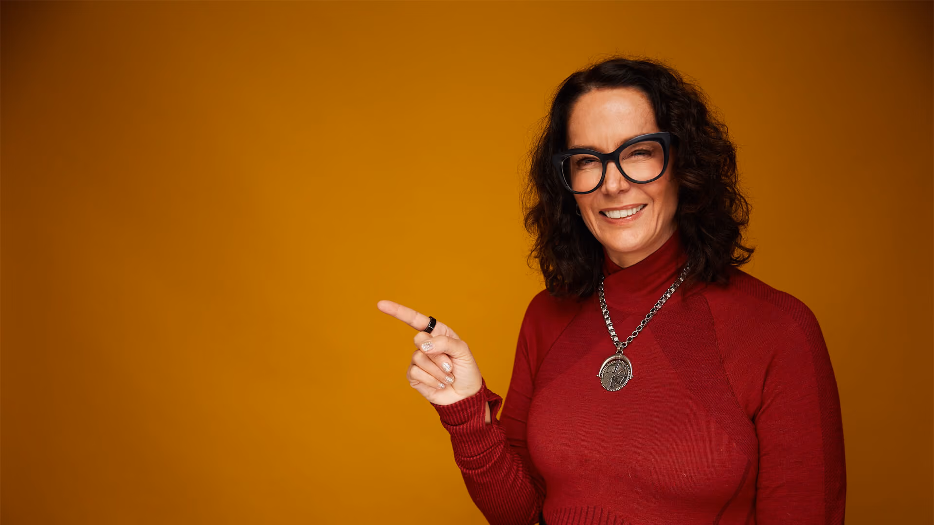 Smiling woman with curly hair and glasses in red sweater pointing left on orange background.