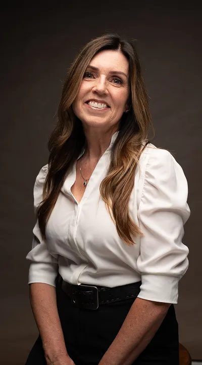 Smiling woman with long brown hair wearing a white blouse and black pants against a dark background.