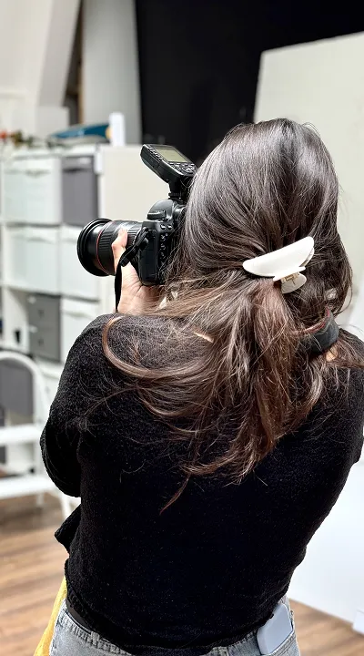 Photographer with dark hair and white clip, holding a professional camera with flash, side view from behind.