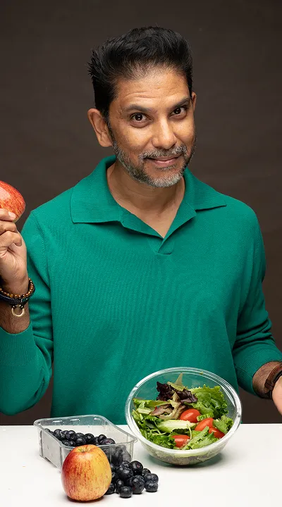 Man in green shirt holding an apple with a salad bowl and grapes on the table in front of him.