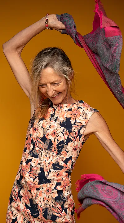 Smiling woman in floral dress holding and waving a patterned scarf against an orange background.
