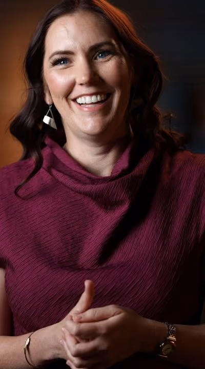 Smiling woman with wavy brown hair wearing a purple top and silver earrings.