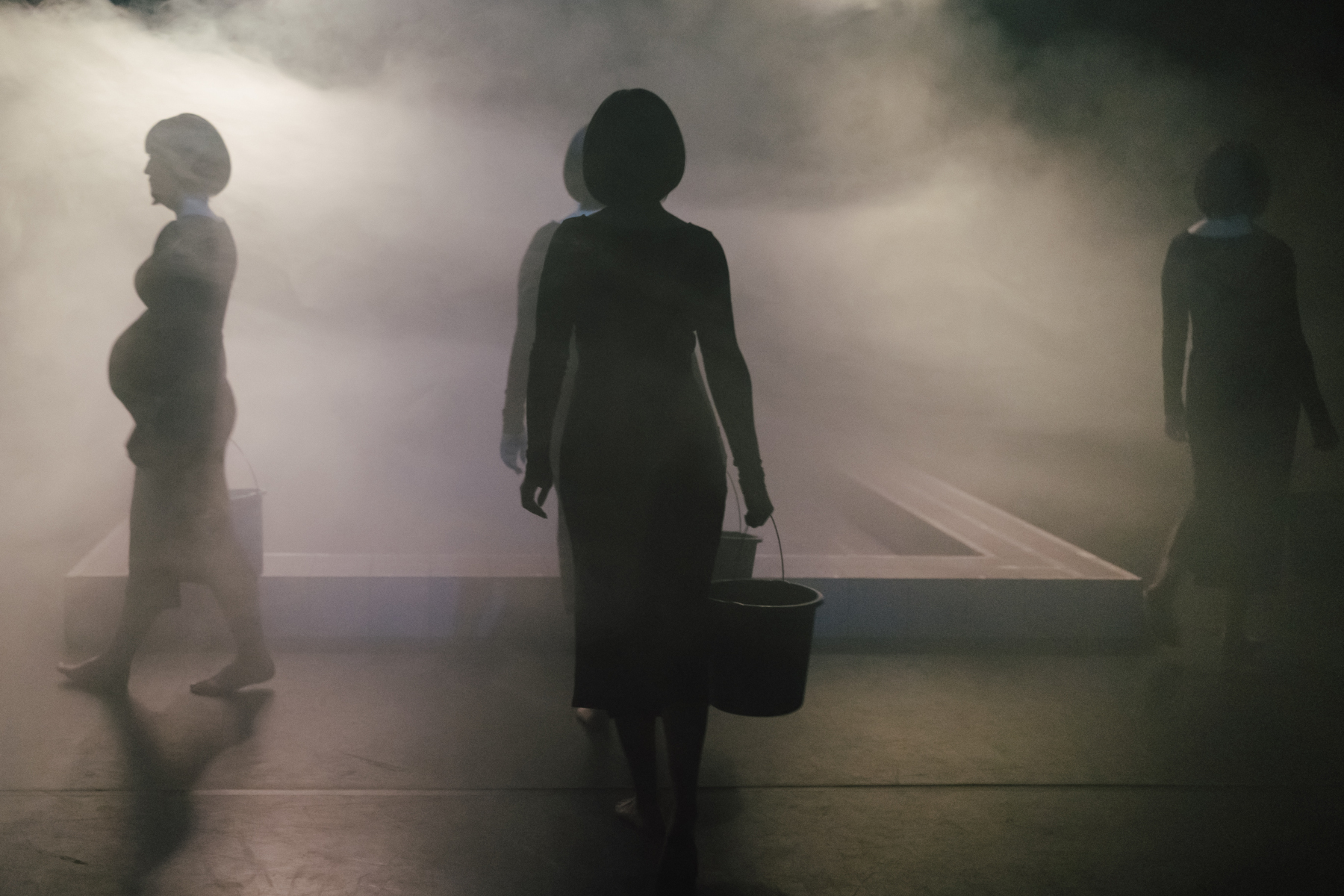 vor Nebel sind als Silhouetten Schwangere in schwarzen Kleidern zu sehen. Sie tragen je einen Eimer.