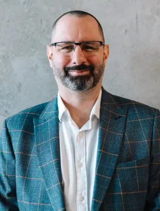 Smiling man with glasses, beard, and short hair wearing a checkered blazer and white shirt against a grey background.