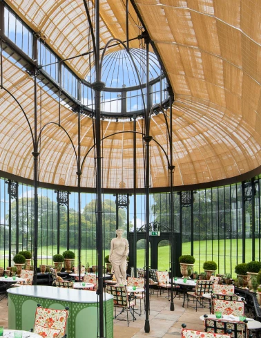 Glass conservatory with a domed ceiling, black metal framing, floral cushioned chairs, and a white statue inside.
