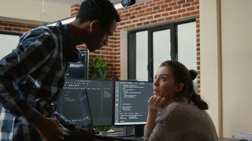 Two software developers, a man and a woman, are discussing code on their computer monitors.