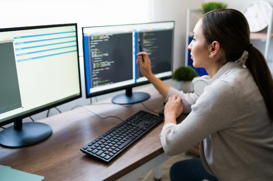Kobieta siedzi przy biurku i analizuje kod na dwóch monitorach, trzymając długopis i wskazując na ekran.