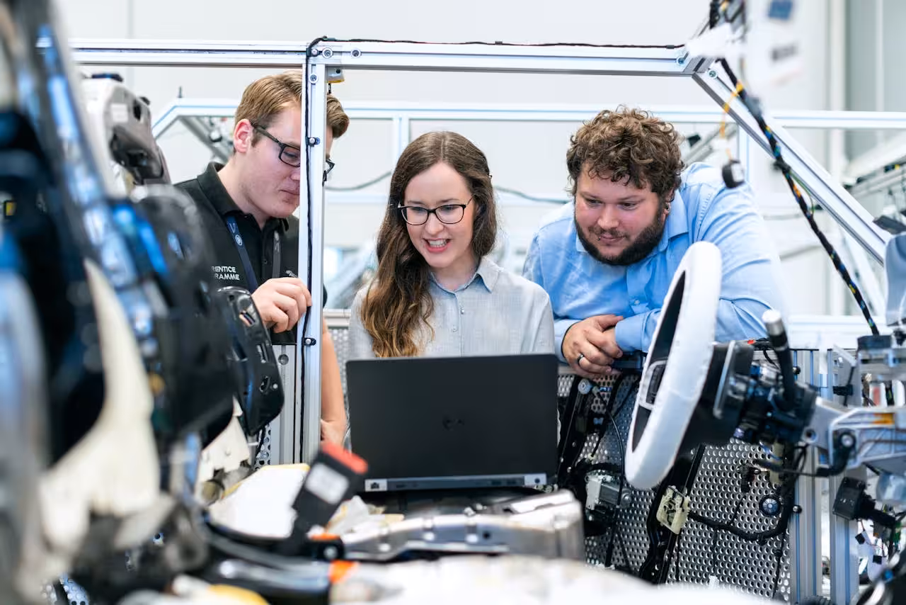Three focused engineers reviewing code on a laptop amidst complex industrial machinery, suggesting work on AI-driven Business Process Automation (BPA) for manufacturing or R&D processes.