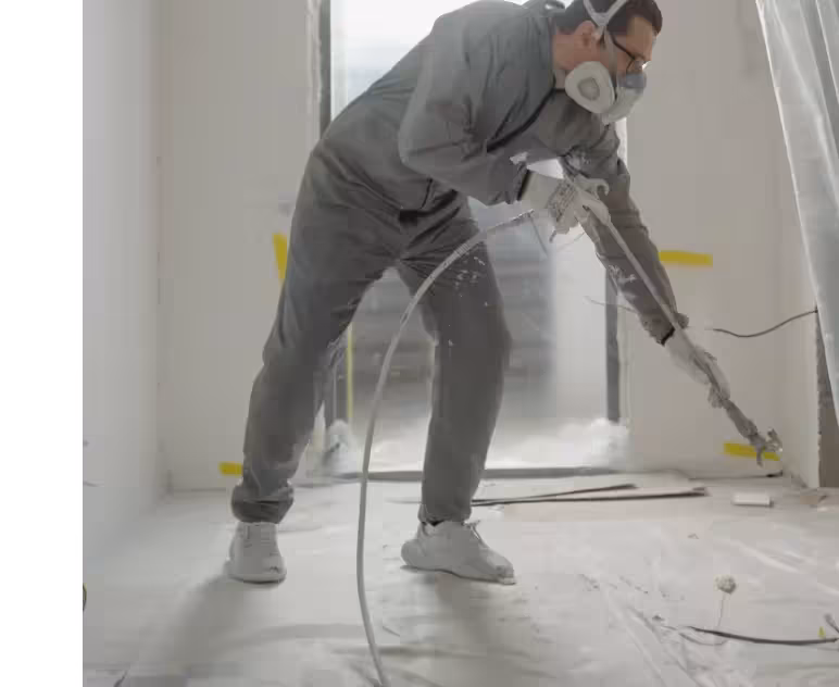 A construction worker wearing gray protective overalls, a mask, and goggles sprays white paint or construction chemicals in a modern, empty room using a professional spray gun. The room is partially covered with plastic sheeting.