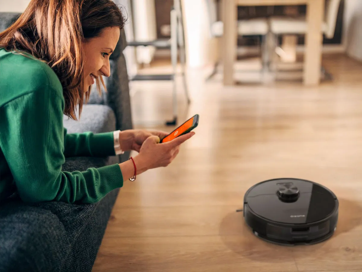 Woman in green sweater lying on couch controlling robot vacuum with smartphone on wooden floor.