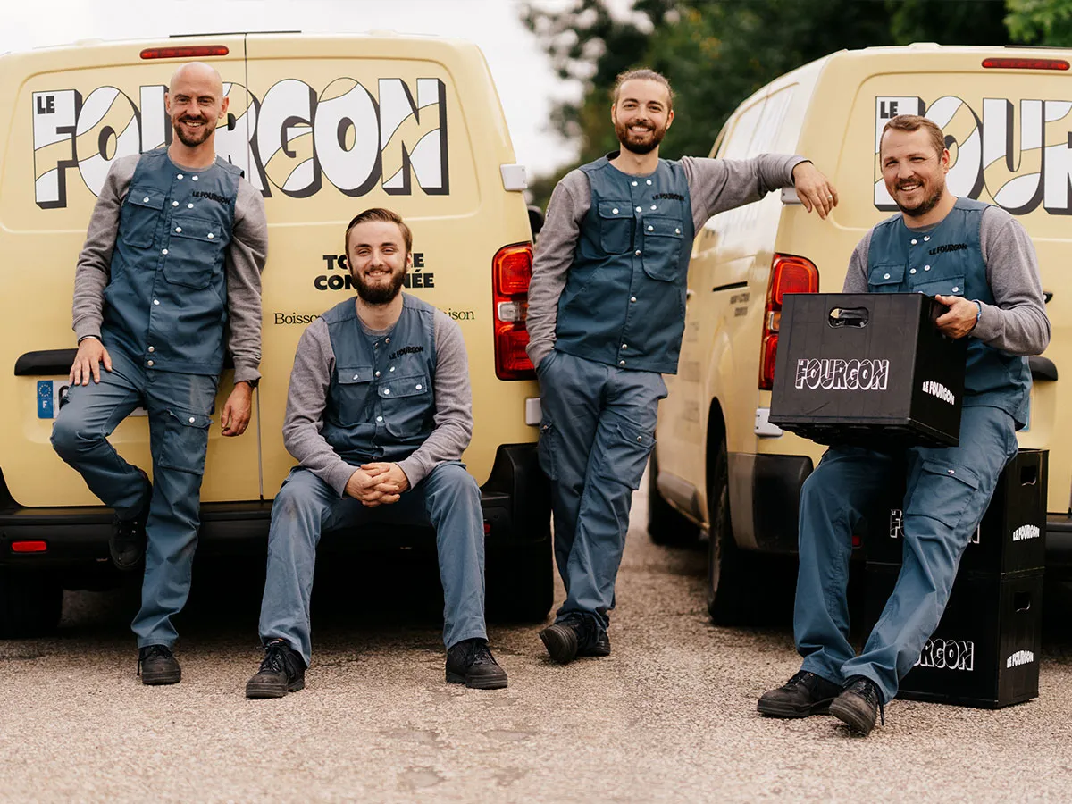 Four men in matching work uniforms pose by yellow vans with 'Le Fourgon' logo and holding branded crates.