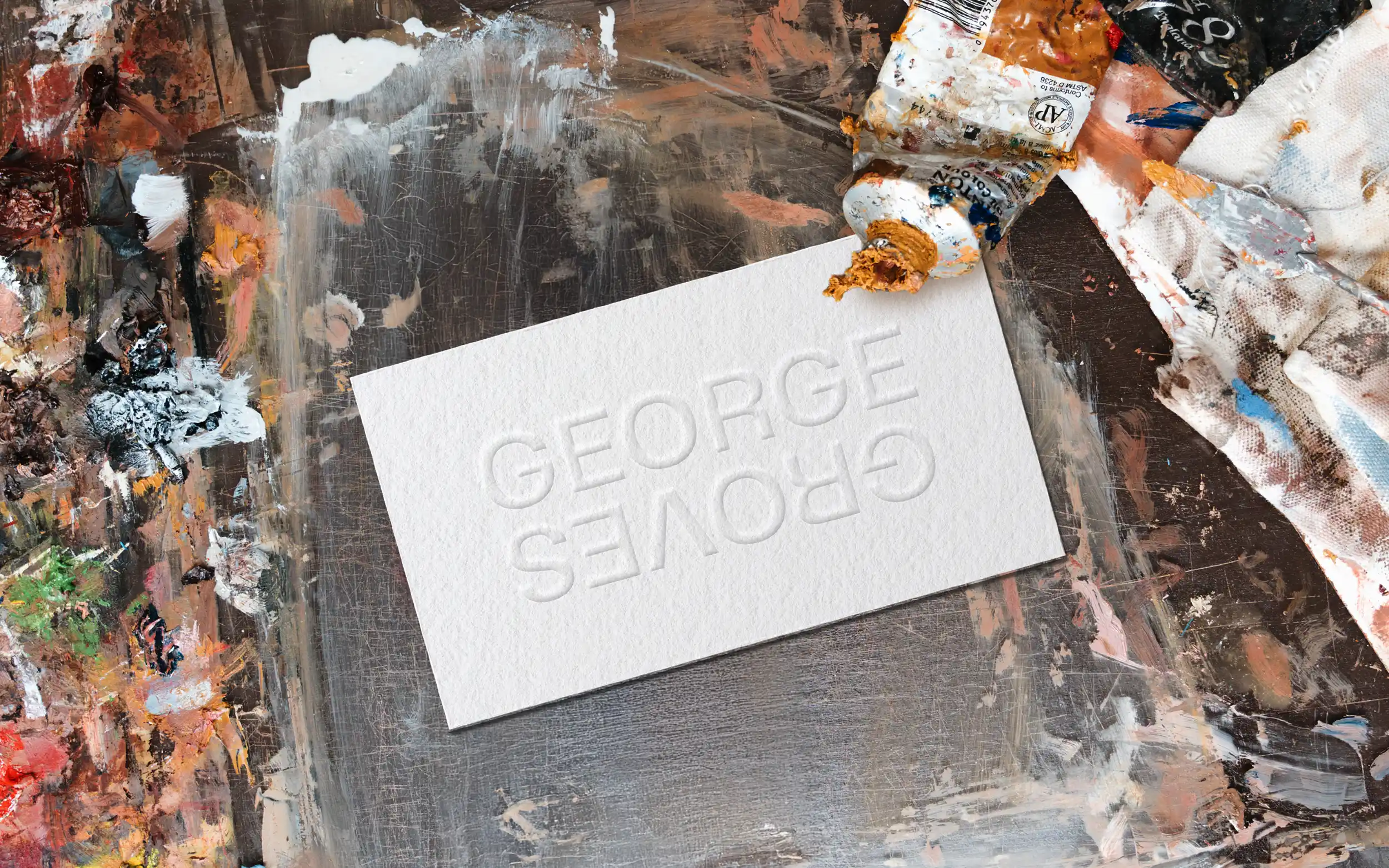 White business card with embossed 'George Groves' branding, placed on a tabled covered in paint marks.