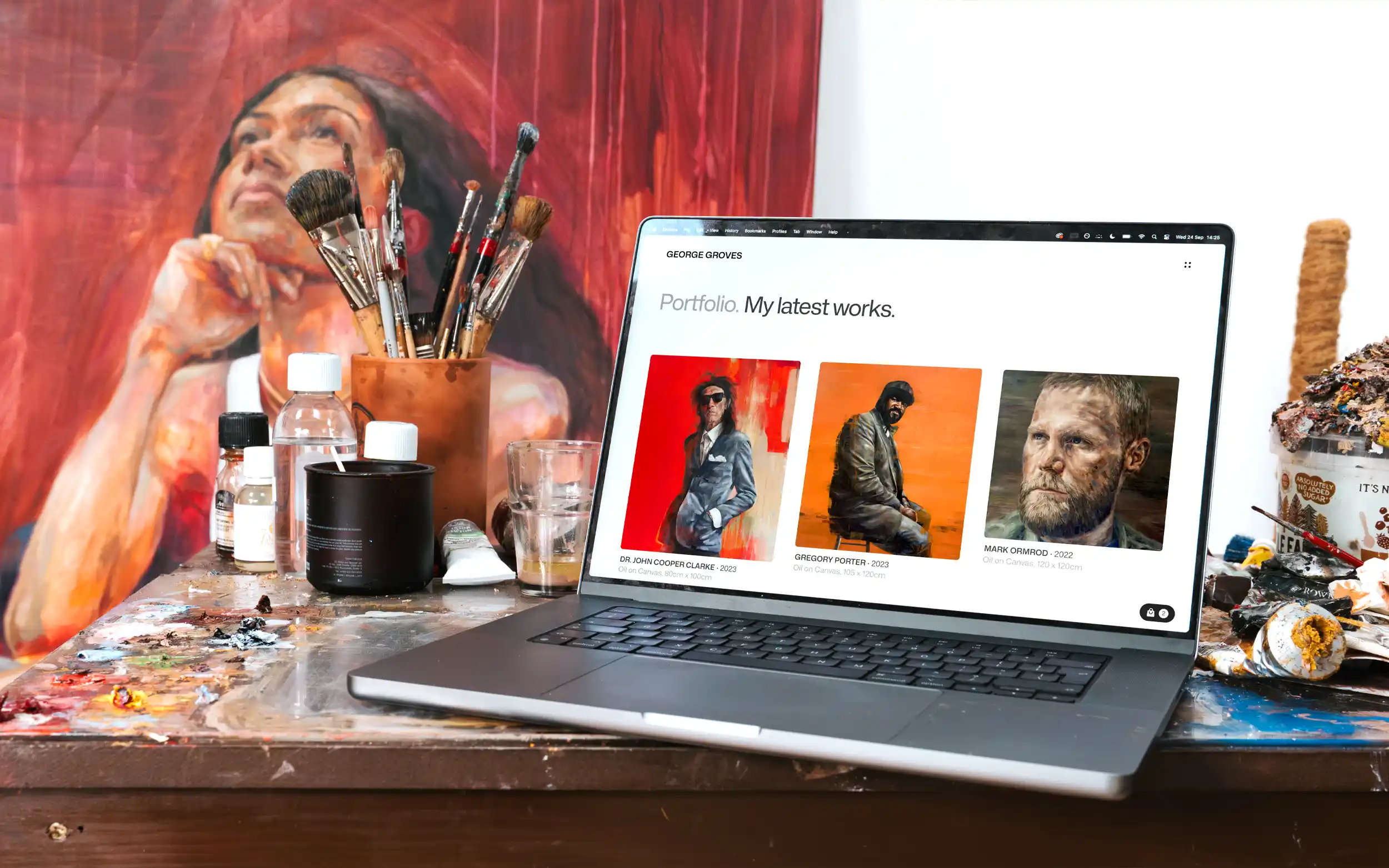 Laptop displaying the George Groves website portfolio page, placed on an artist work station, surround by artist painting materials.