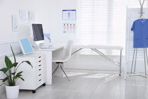 Bright doctor office with white desk, computer, examination table.
