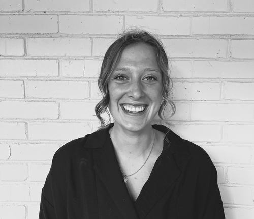 Portrait of Cadence Cutler — Young woman standing in front of a white brick wall, smiling.