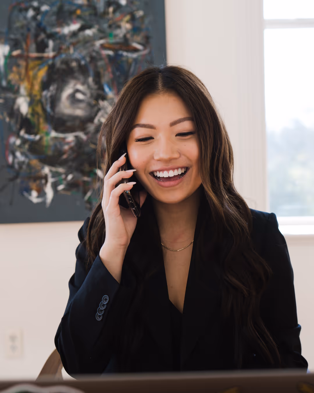 Woman in a suit, standing behind a reception desk, on the phone,  looking at a computer screen pleasantly. 