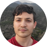 Hombre joven con cabello oscuro rizado, camiseta roja, con fondo de árboles y montañas difusas.
