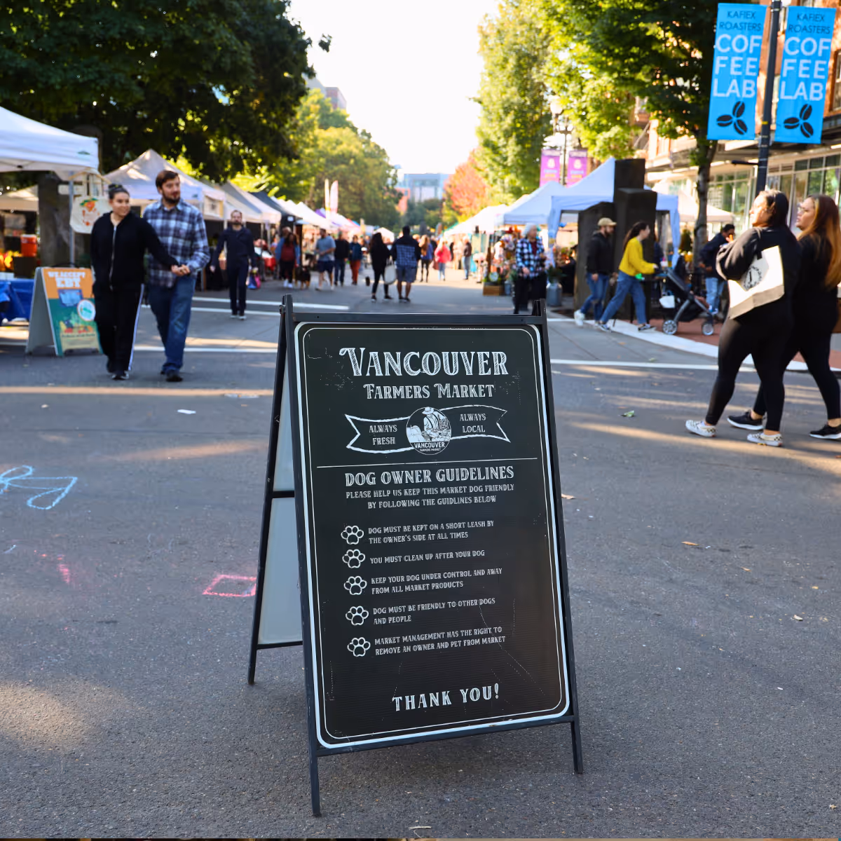  A sidewalk sign at Vancouver Farmers Market lists dog owner guidelines, with market stalls and people in the background.