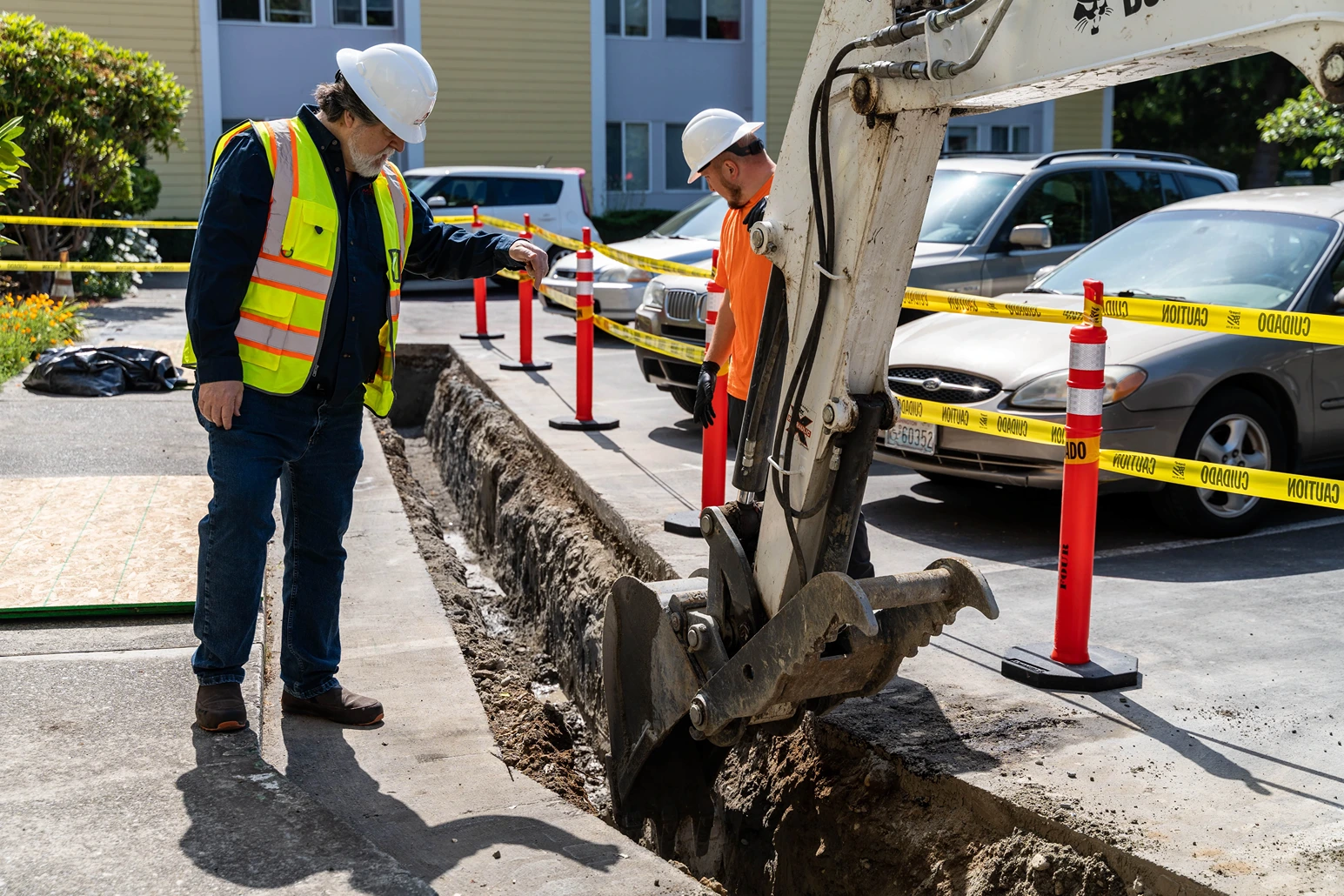 Supervisor in a high-vis safety vest pointing down into a paved trench next to a white excavator bucket.