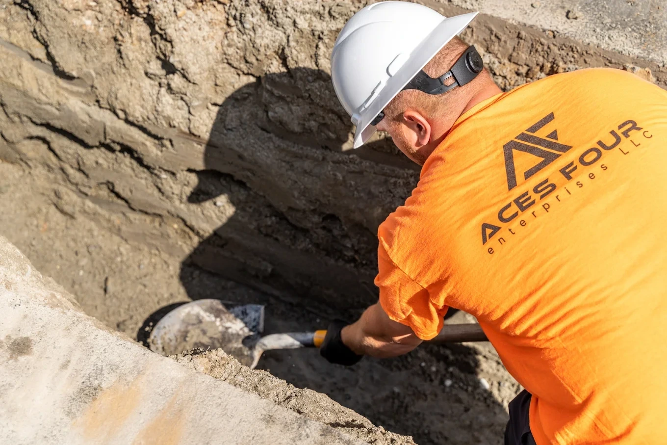 Construction worker in a hard hat and orange shirt digging with a shovel inside a trench.