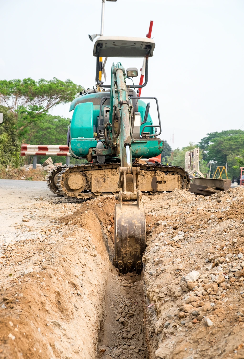 Compact teal excavator digging a narrow trench along a dirt road for sewer line installation.
