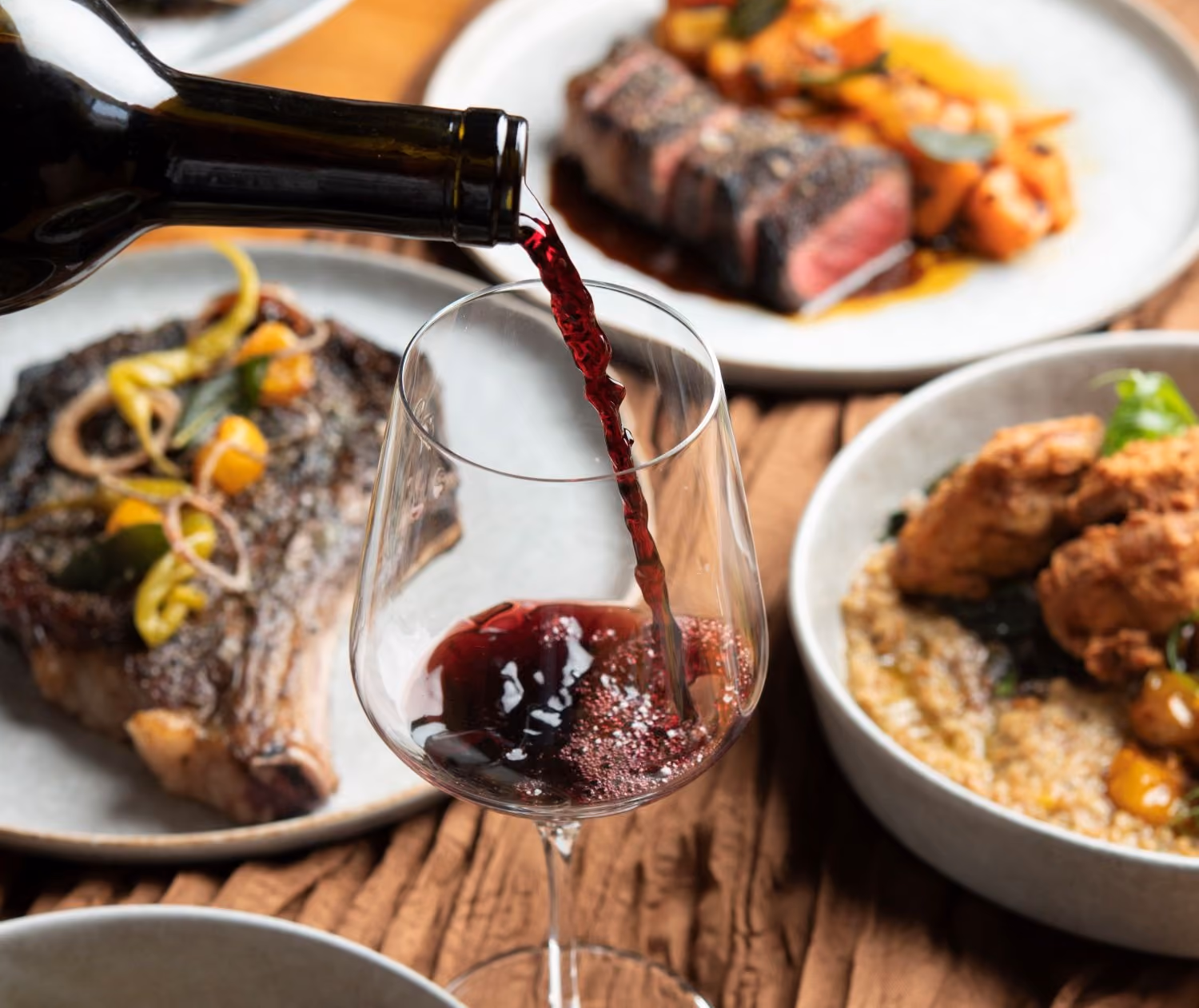 Glass of red wine being poured with various dishes of food including a steak