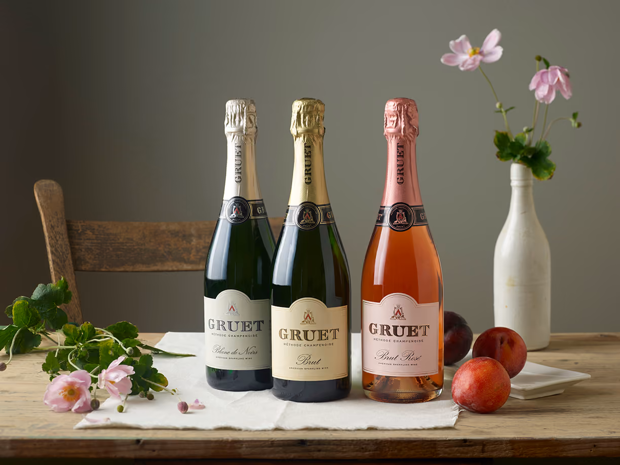Three bottles of Gruet wine lined up on a table with  a vase of flowers and some nectarines