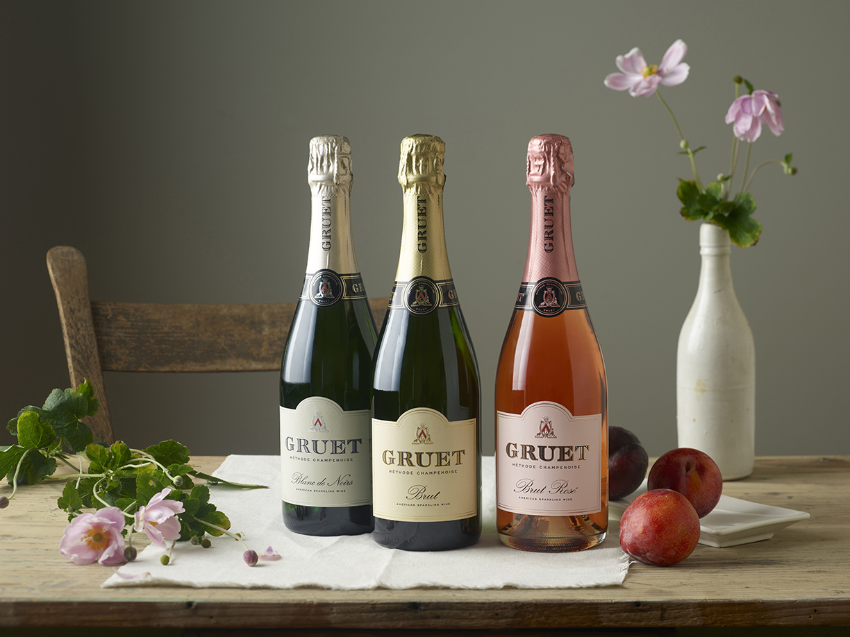 Three bottles of Gruet wine lined up on a table with  a vase of flowers and some nectarines