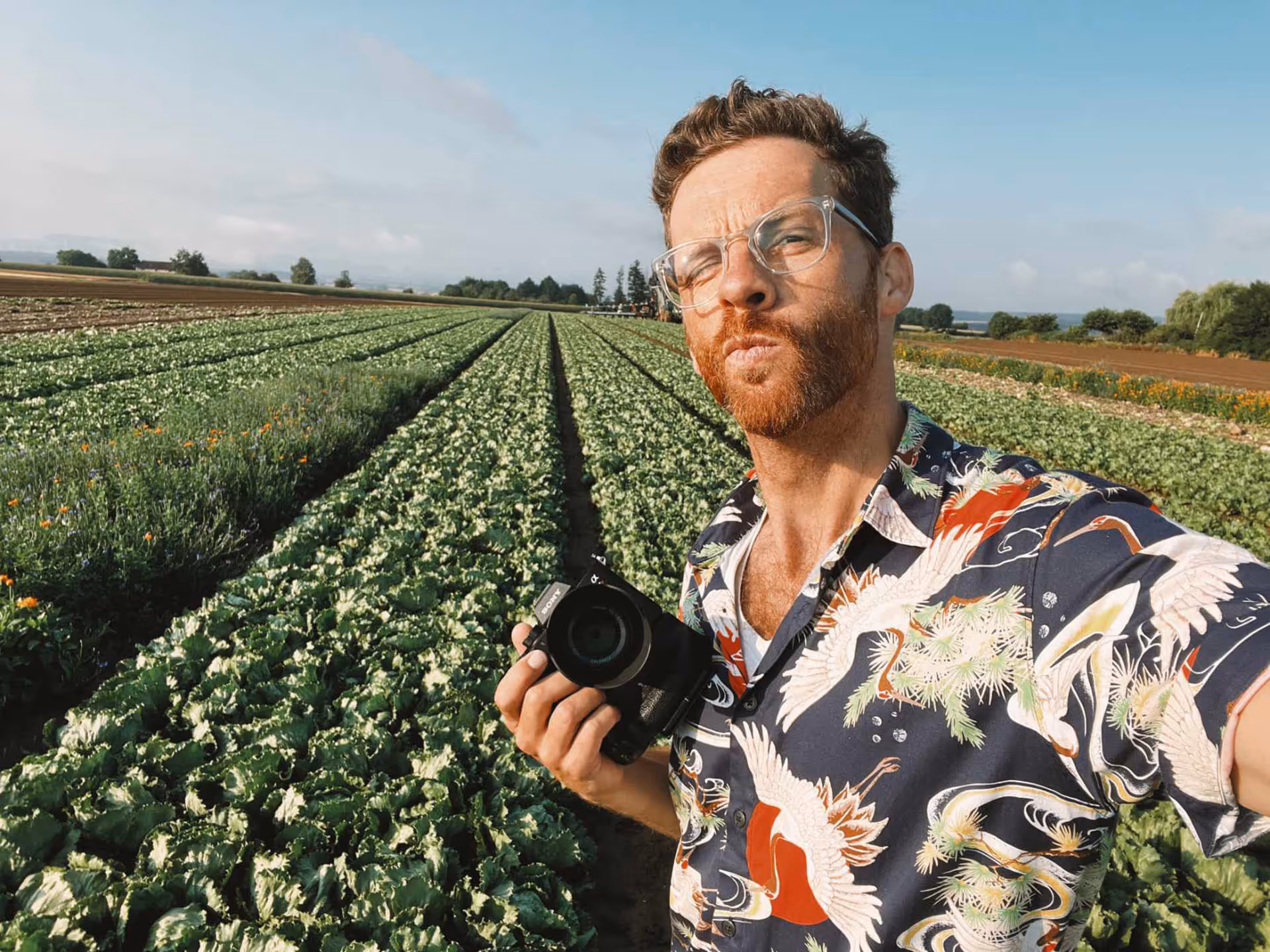 Mann mit Kamera in der Hand macht Selfie auf einem Feld mit grünem Gemüse unter blauem Himmel.