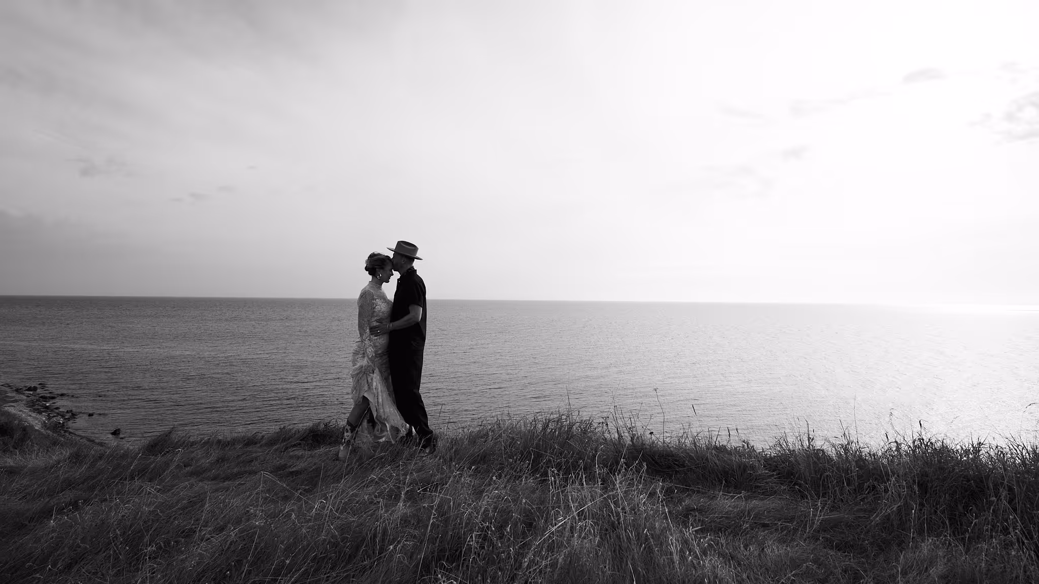 Paar steht am Rand einer Klippe vor dem Meer, der Mann küsst die Stirn der Frau, Schwarzweißaufnahme.