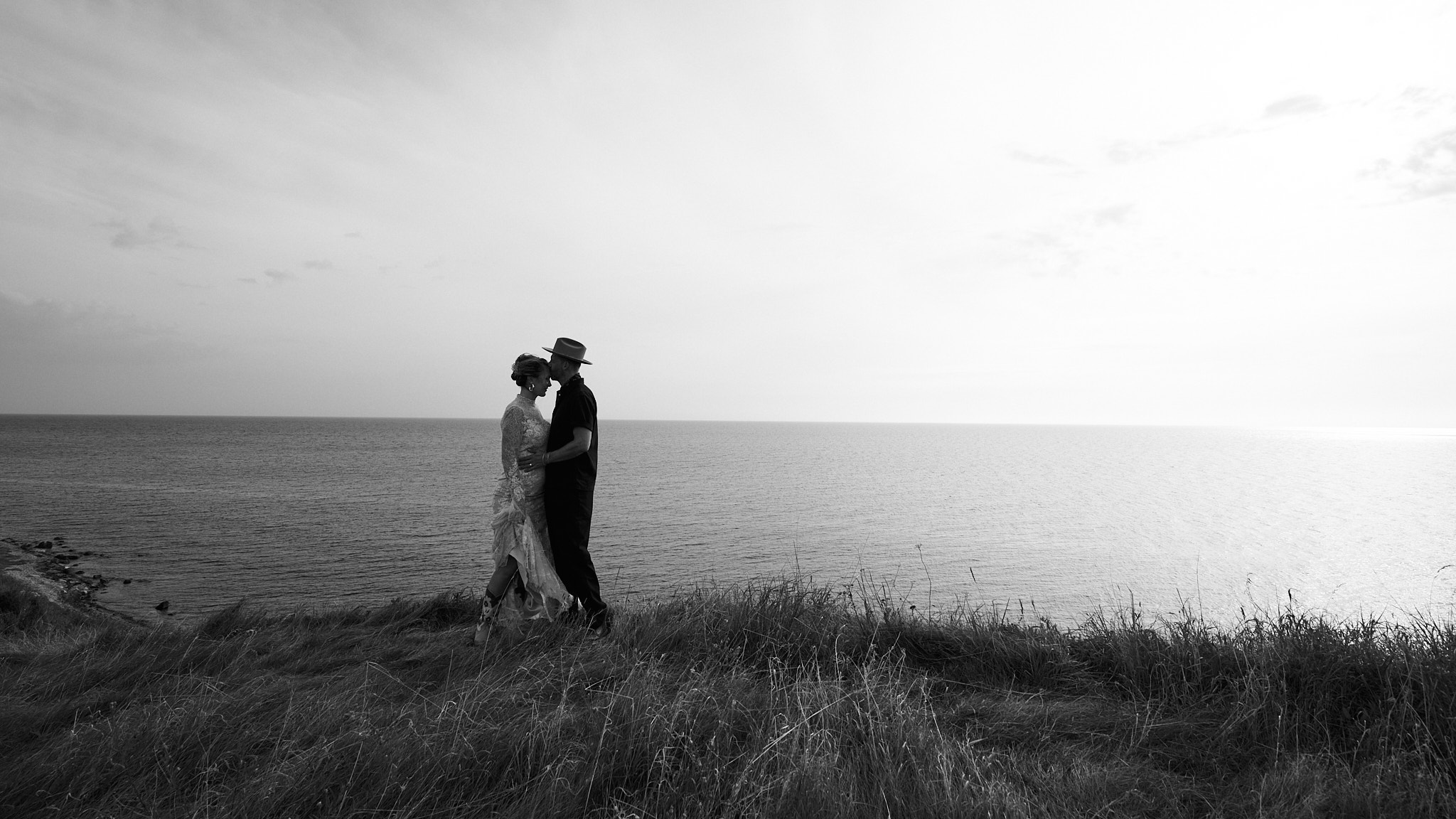 Paar steht am Rand einer Klippe vor dem Meer, der Mann küsst die Stirn der Frau, Schwarzweißaufnahme.