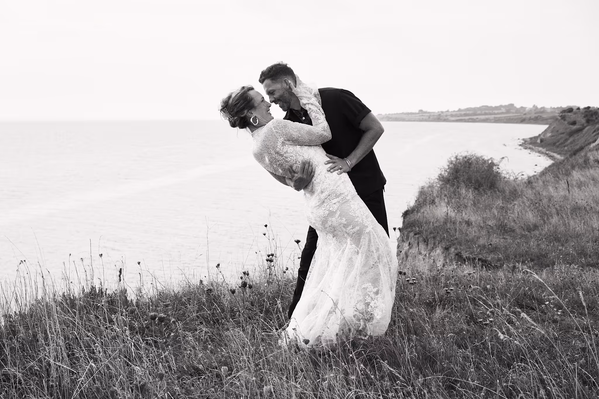 Ein Paar in Hochzeitskleidung lacht und tanzt am Rand einer Klippe über dem Wasser.