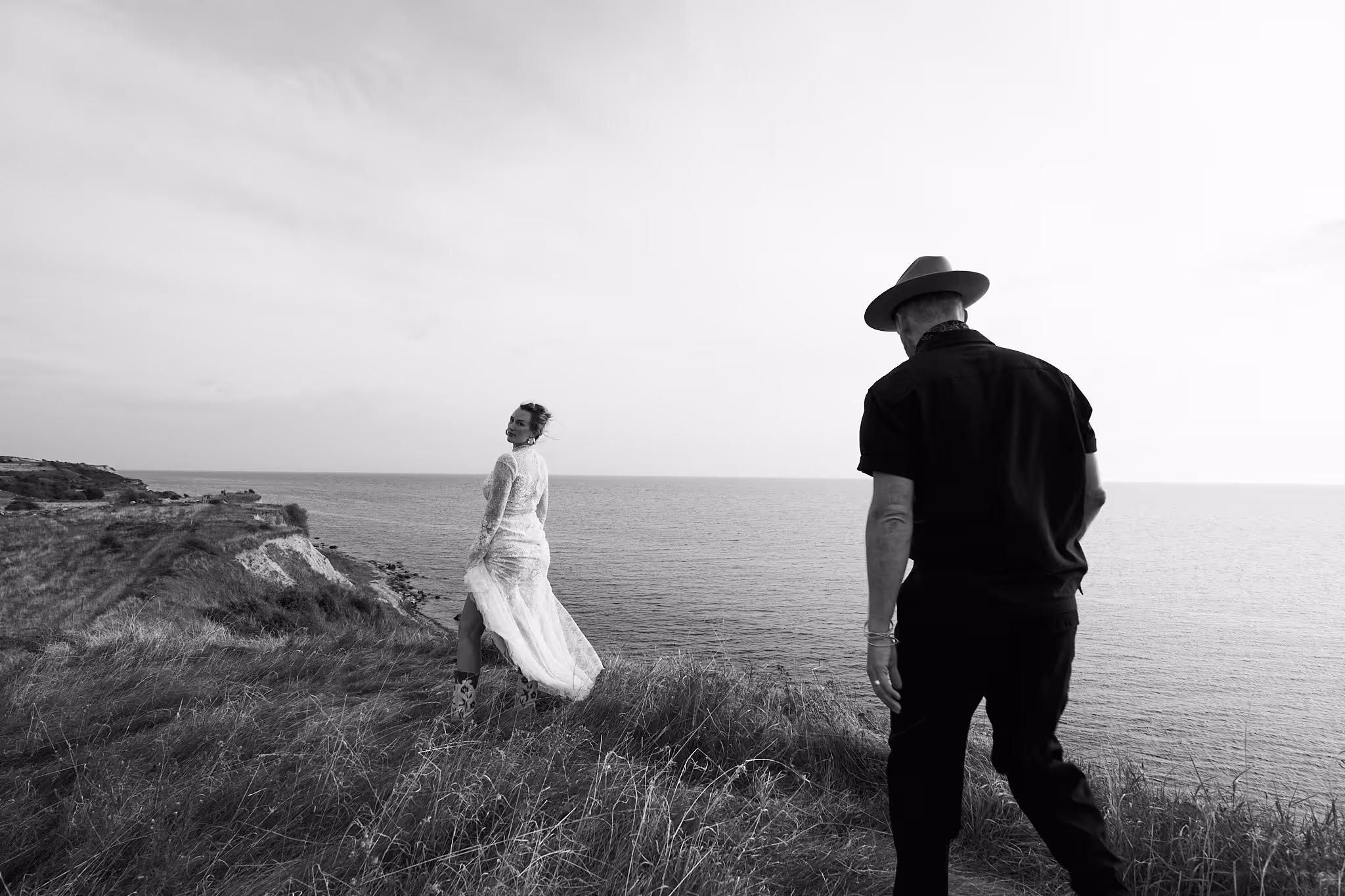 Ein Mann mit Hut und eine Frau in einem weißen Spitzenkleid stehen auf einer Klippe mit Blick auf das Meer.
