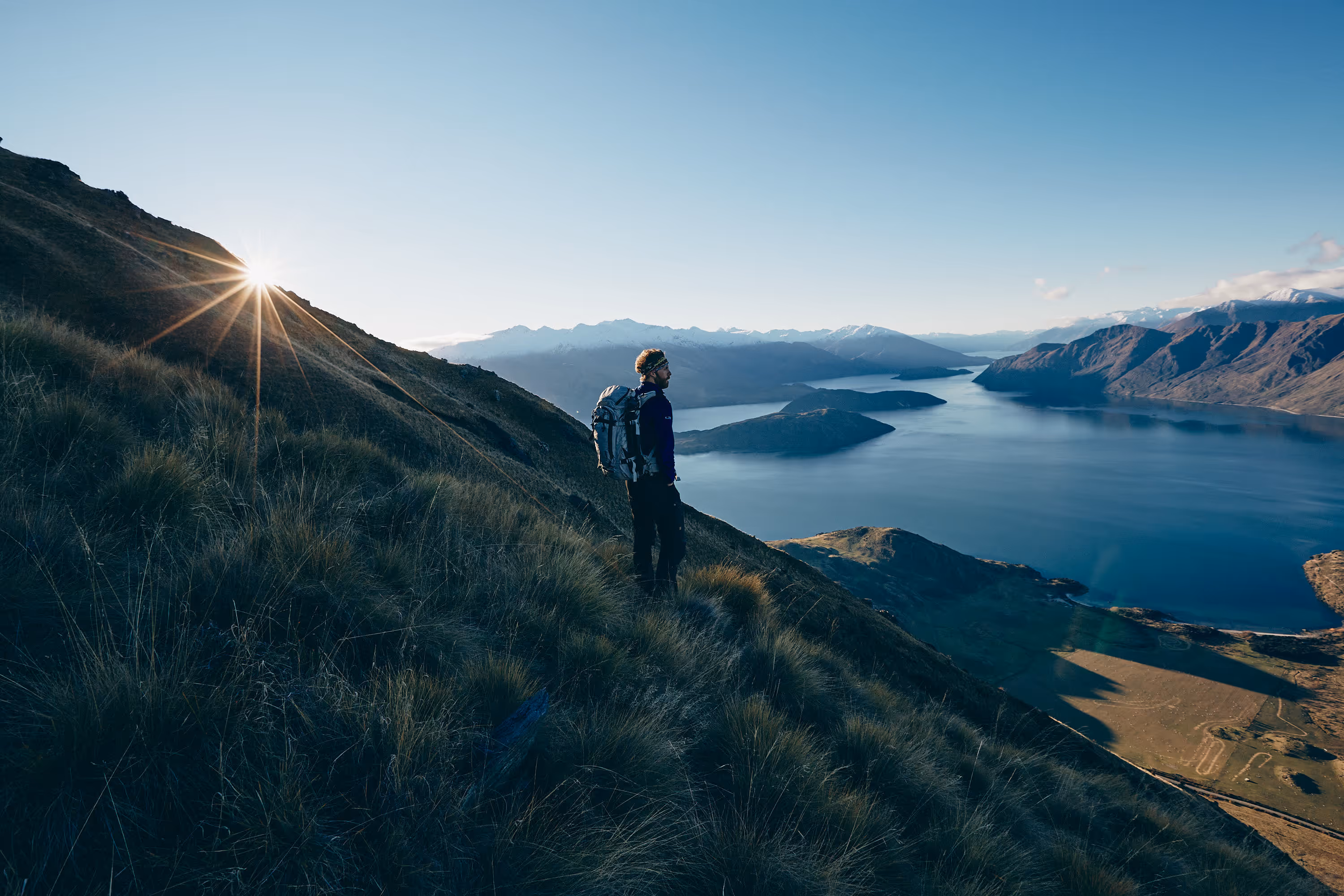 Wanderer steht auf einem grasbewachsenen Hang und blickt auf einen See und Berge bei Sonnenaufgang.