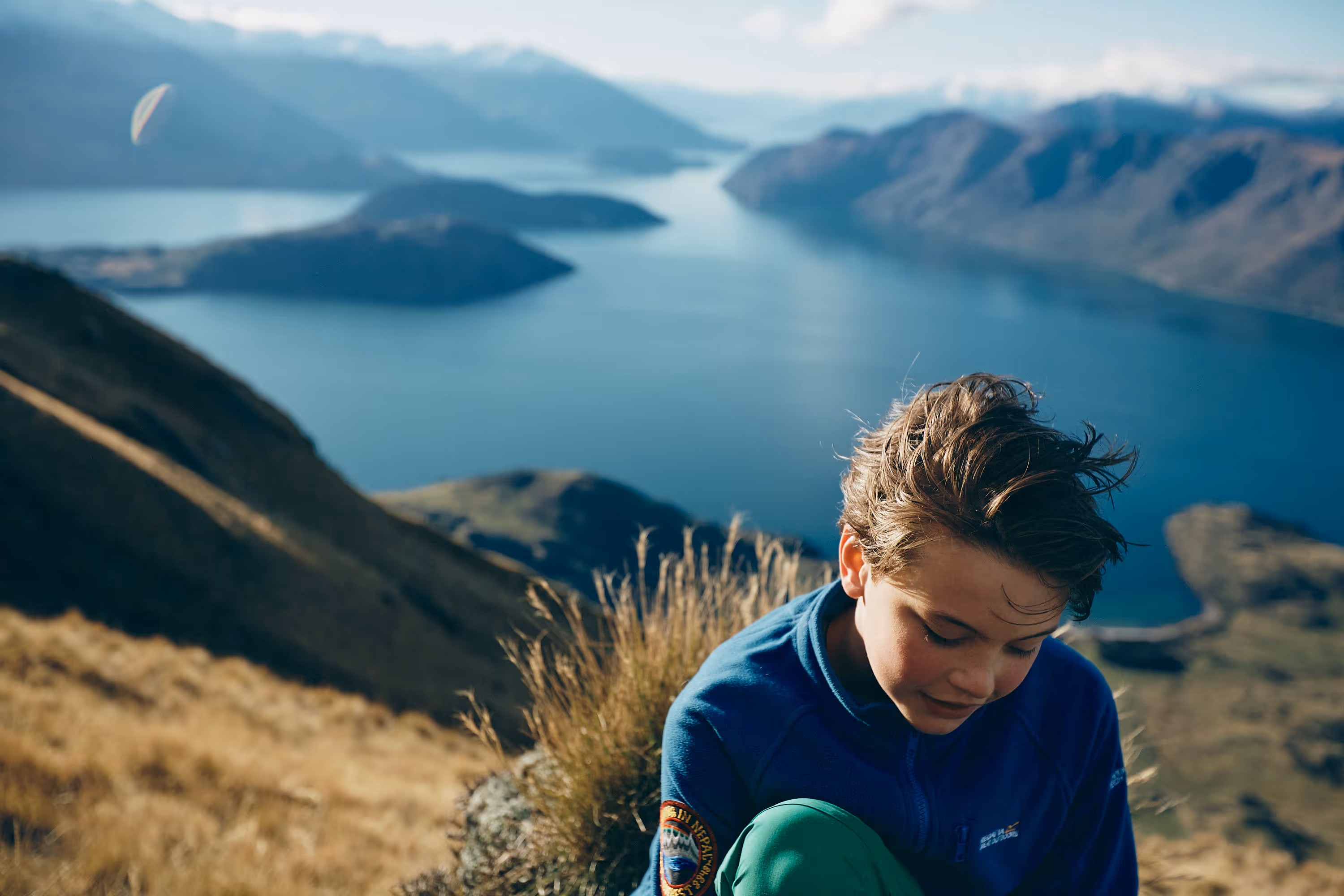 Junge mit blauem Pullover sitzt auf einem felsigen Hügel mit Blick auf einen See und Berge im Hintergrund.