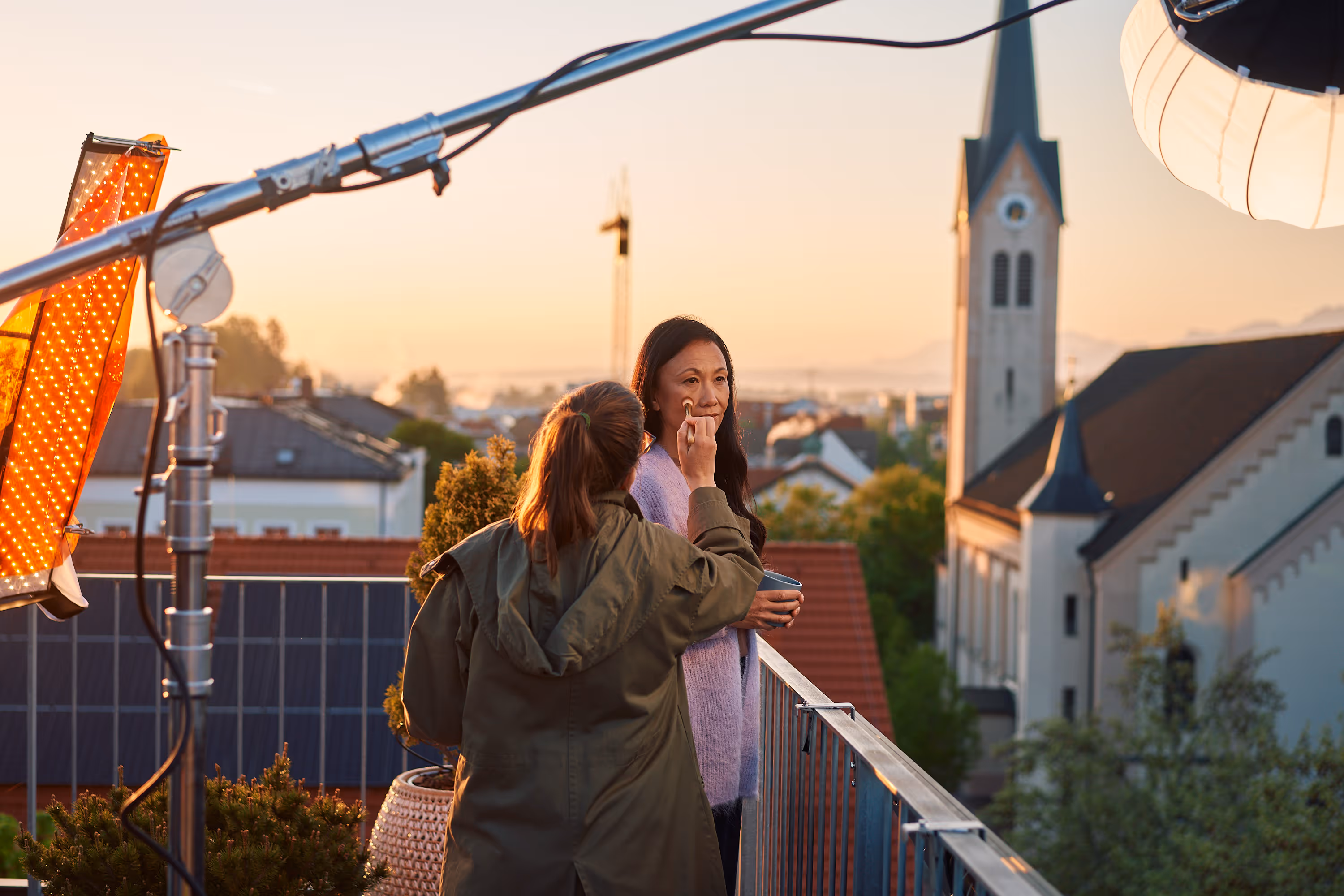 Eine Frau bekommt auf einem Balkon bei Sonnenaufgang professionelles Make-up aufgetragen, mit Stadtansicht und Kirche im Hintergrund.