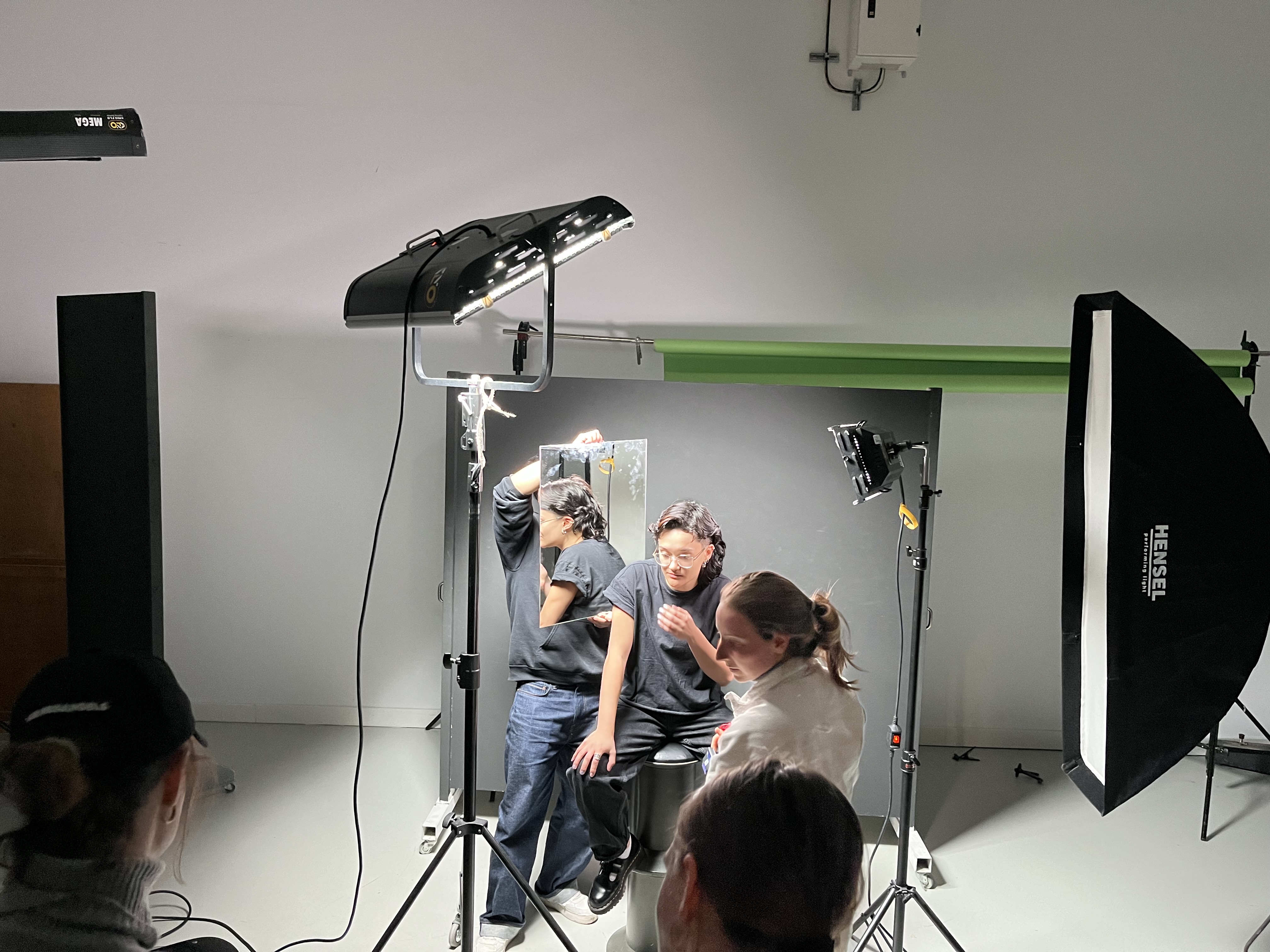 Fotostudio mit zwei Personen, eine sitzt auf einem Hocker, die andere steht und hält einen Spiegel, um das Gesicht der sitzenden Person zu reflektieren.