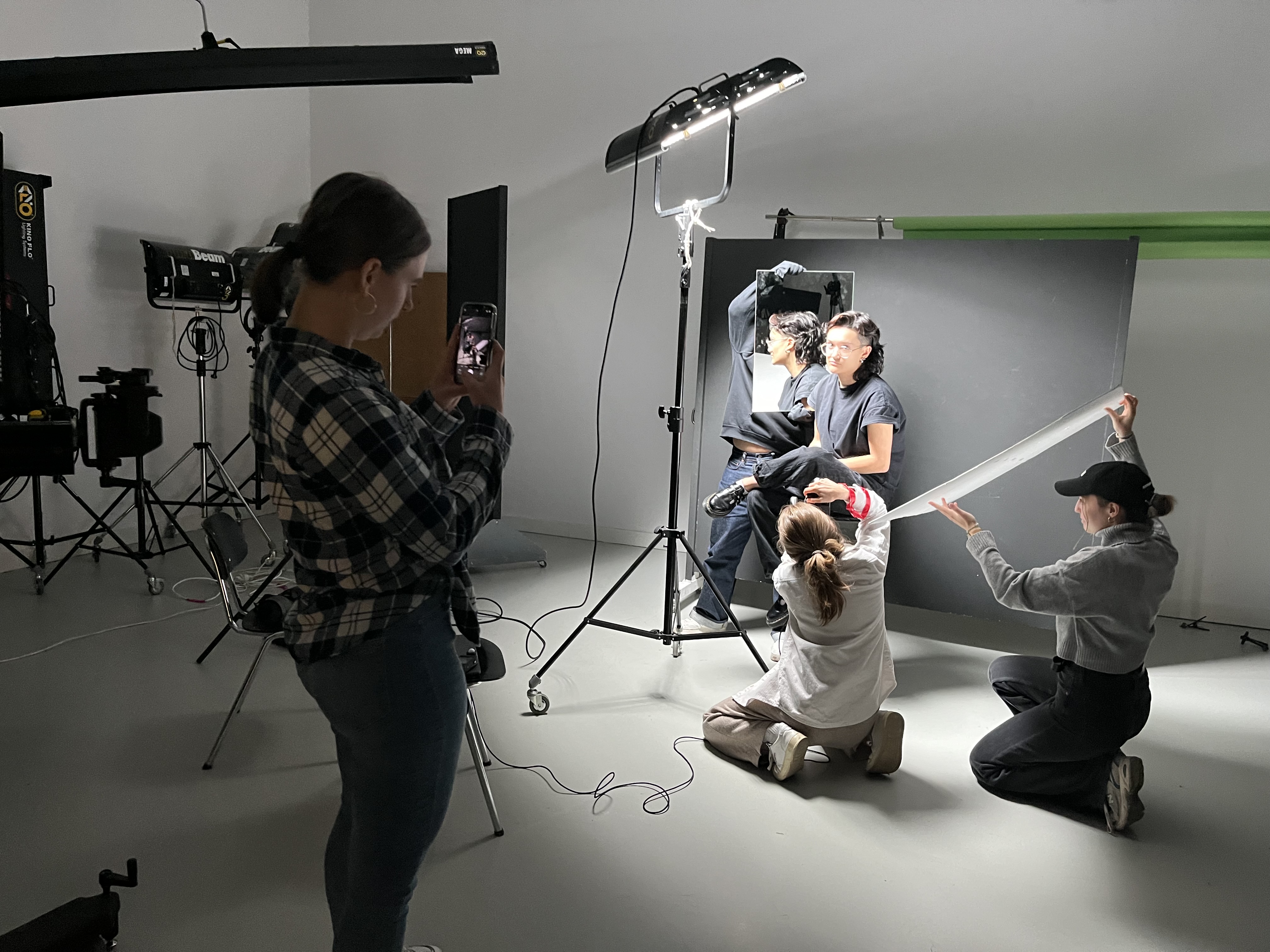 Fünf Personen in einem Fotostudio: eine Person fotografiert mit dem Handy, zwei knien und arrangieren ein weißes Lichtreflektorband, zwei sitzen vor einer schwarzen Wand mit Spiegel im Hintergrund.
