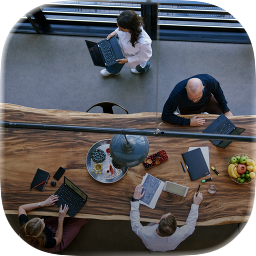 Vier Personen arbeiten an einem langen, rustikalen Holztisch mit Laptops, Dokumenten und Obstschale.