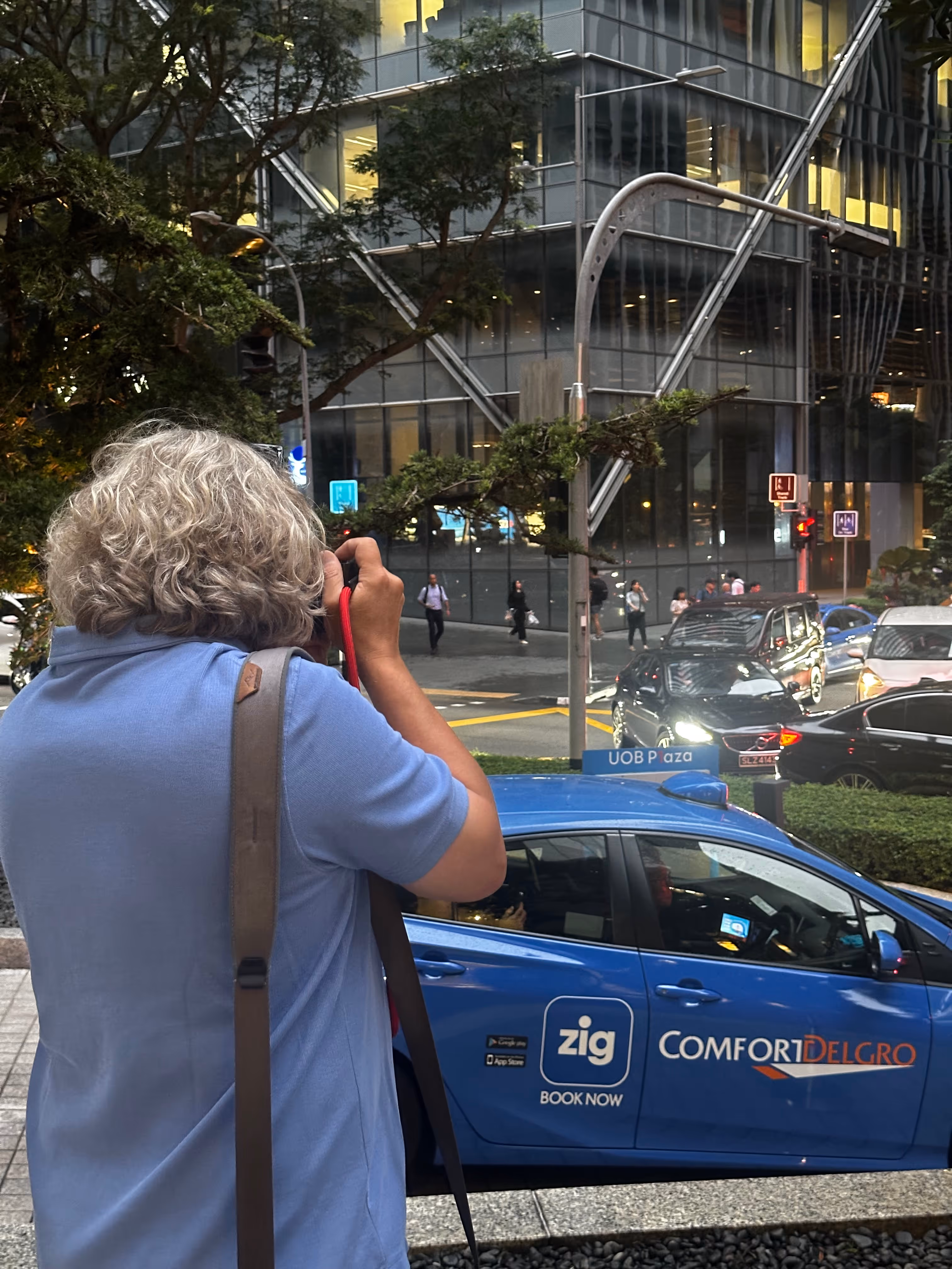 Person mit grauen, lockigen Haaren fotografiert auf einer Straße mit einem blauen ComfortDelGro Taxi und modernen Glasgebäuden im Hintergrund.