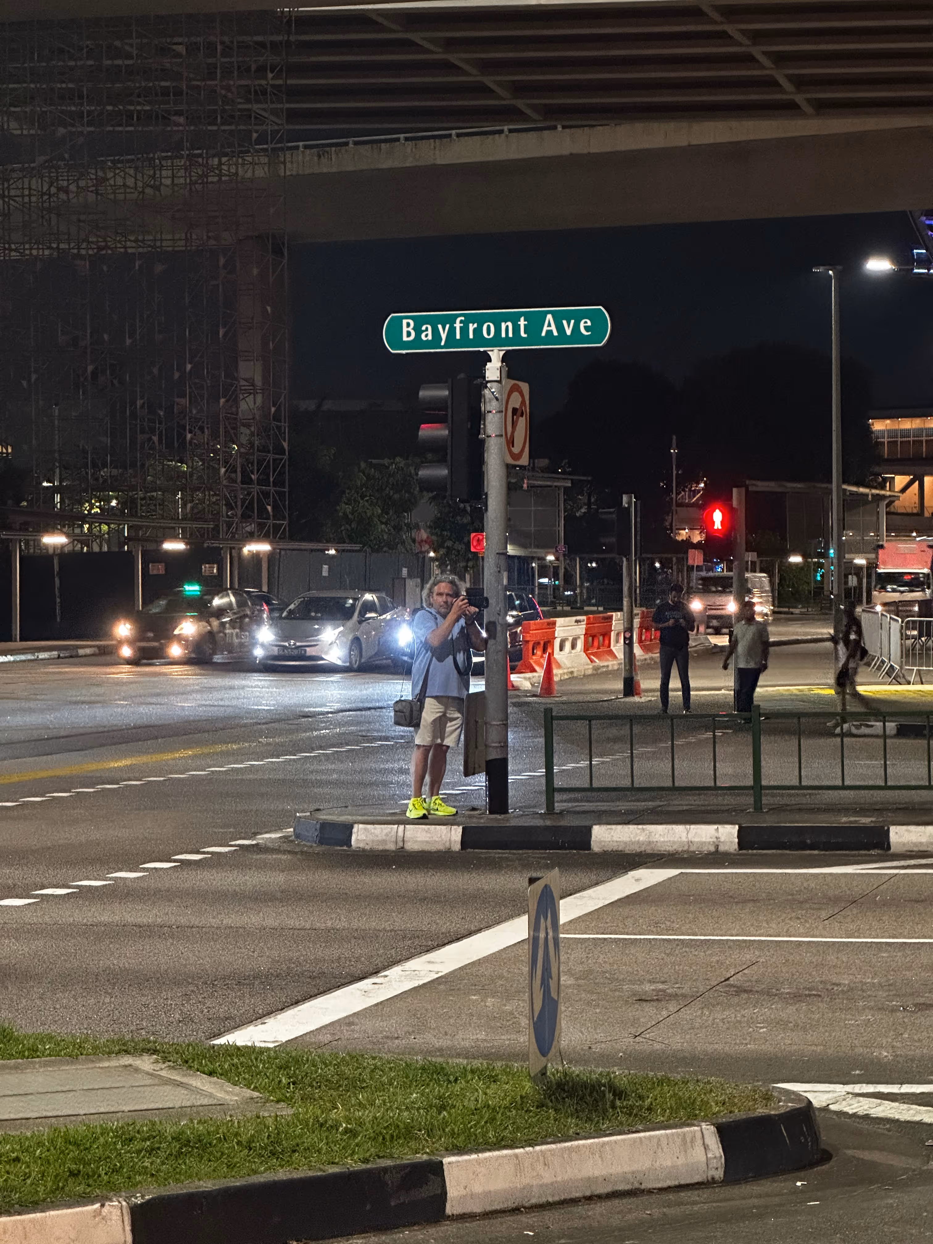 Ein Mann mit neonfarbenen Schuhen steht nachts an einer Kreuzung unter einem Straßenschild mit der Aufschrift Bayfront Ave und fotografiert.
