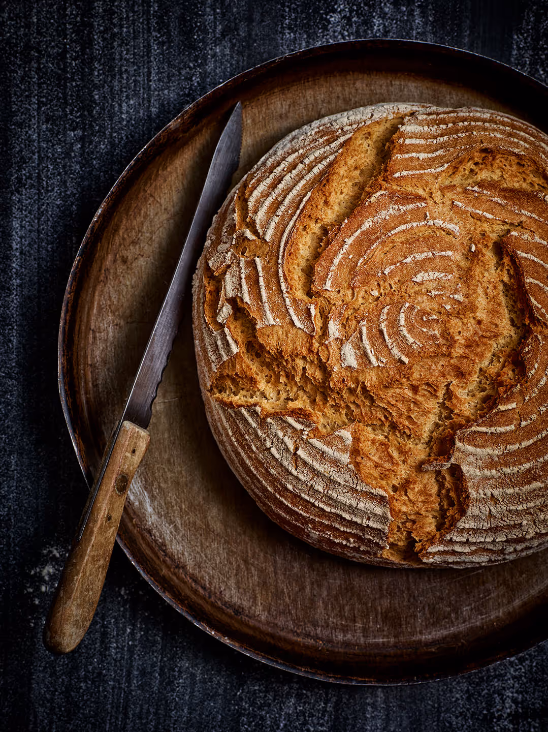 Rundes, rustikales Sauerteigbrot mit einem Muster aus Mehl, auf einem Holzbrett mit einem Brotmesser.